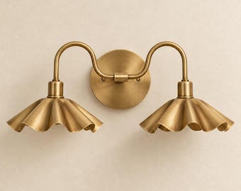Può includere: Una lampada da parete doppia con finitura in ottone spazzolato. È dotata di una piastra circolare con due bracci curvi che si estendono verso l'esterno, ciascuno dei quali sostiene un ampio paralume rivolto verso il basso con bordo ondulato.
