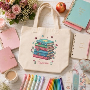 May include: A beige canvas tote bag featuring a central graphic of a stack of five colorful books adorned with pink roses and small butterflies. The name Danielle is printed in pink script below the books. The bag has two sturdy shoulder straps.