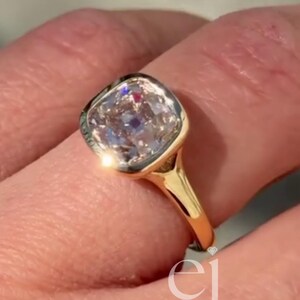 Peut inclure: Une bague en or avec une pierre centrale taille coussin montée dans un serti clos lisse. L'anneau est en or jaune poli et la pierre présente des facettes claires qui reflètent la lumière. Les lettres ej sont visibles en bas à droite.