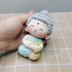 May include: A small crocheted figurine of a seated figure with a grey textured hairstyle, a red dot on the forehead, and pink cheeks. The figure wears a pale yellow and light blue garment, featuring a smiling expression with closed eyes.