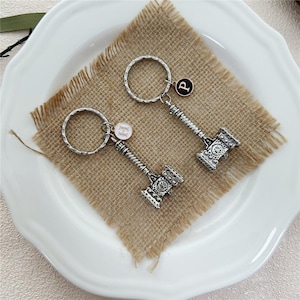Peut inclure: Deux porte-clés en métal argenté en forme de marteau avec des manches texturés et des têtes détaillées. Chaque porte-clés comprend un anneau circulaire et une petite breloque ronde, l'une avec la lettre H en blanc et l'autre avec la lettre P en noir.