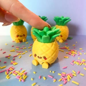 May include: A collection of small, yellow pineapple-shaped erasers with green leafy tops and smiling faces. Each pineapple has two small yellow feet at the base. The erasers are scattered among small, colorful pink and yellow sprinkles.