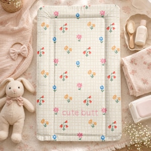 Può includere: Un fasciatoio rettangolare con sfondo color crema, un sottile motivo a griglia e una stampa floreale ripetuta nei toni del rosa, blu e arancione. Il testo "cute butt" è ricamato con filo rosa nella parte inferiore centrale.