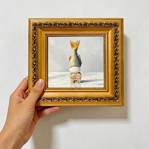 May include: A square oil painting in an ornate gold frame depicting a vertical stack consisting of a fish tail pointing upward, a white egg in the centre, and a cross-section of a fish at the base, all set against a light grey background.