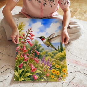 Könnte beinhalten: Ein rechteckiger Kunstdruck mit einem lebendigen Aquarellgemälde eines Kolibris mit roter Kehle, der neben hohen rosa Fingerhutblumen schwebt. Der Hintergrund zeigt eine üppige grüne Landschaft unter einem hellblauen Himmel mit Wolken.