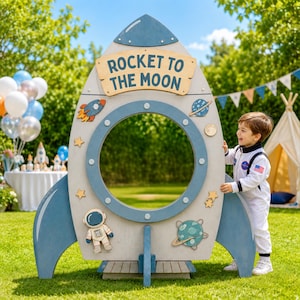 May include: A large, light gray and blue wooden rocket ship photo cutout stand with a circular opening. The sign features the text ROCKET TO THE MOON, along with decorative wooden cutouts of a small rocket, an astronaut, and planets.