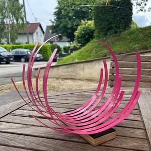 May include: A decorative sculpture featuring multiple curved, bright pink metal strips arranged in a fan-like, wave pattern. The strips are anchored into a rectangular wooden base with a black top surface, creating a dynamic, rhythmic visual effect.