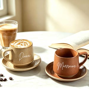 Op de afbeelding: Twee keramische mokken met bijpassende schoteltjes, één beige en één terracotta, beide met een gespikkelde afwerking en een naam in witte schrijfletters. De beige mok is gelabeld met Diana en bevat koffie, terwijl de terracotta mok is gelabeld met Marraine.