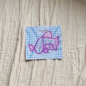 May include: A square packaging card featuring a blue and white gingham pattern with a pink outline illustration of a betta fish. The text reads tiredCo Crafts, ROSY BETTA, and PEEL AND STICK WITH STATIC. The fish has detailed fins and scales.