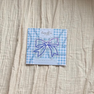 May include: A square packaging card featuring a blue and white gingham pattern with a purple outline of a bow in the center. The text reads tiredCo Crafts, CUTESY BOW SUNCATCHER, and PEEL AND STICK WITH STATIC. The design is clear and centered.
