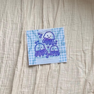 May include: A square sheet of clear static cling stickers featuring three purple jellyfish illustrations with smiling faces and small bubbles. The backing paper has a light blue and white gingham pattern and the text JELLYFISH PALS PEEL AND STICK WITH STATIC.