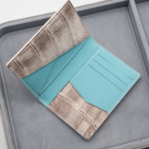 Puede incluir: Una cartera plegable con un exterior de cuero gris con textura de cocodrilo y un interior de color azul turquesa vibrante. El interior incluye varias ranuras para tarjetas y un bolsillo lateral, con costuras blancas en los bordes.