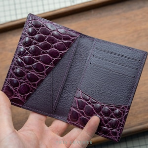 Puede incluir: Una cartera morada con un patrón texturizado de relieve de cocodrilo en los bolsillos exteriores y un acabado granulado en el interior. La cartera incluye tres ranuras horizontales para tarjetas en el lado derecho y el texto TRIS LEATHER abajo.