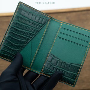 May include: A dark green leather wallet featuring a textured crocodile skin pattern on the pockets and smooth leather on the interior. The wallet includes three card slots and orange contrast stitching. The text TRIS LEATHER is printed at the top.