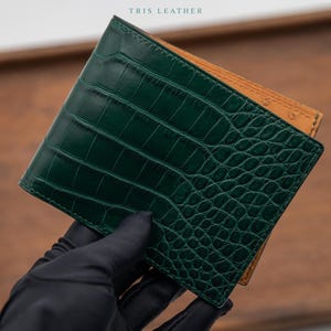 Könnte beinhalten: Ein zweifach faltbares Portemonnaie mit einer dunkelgrünen Außenseite mit Krokodilleder-Struktur und einem hellbraunen Innenbereich. Der Schriftzug TRIS LEATHER ist in kleinen schwarzen Buchstaben oben mittig aufgedruckt. Gehalten in einer schwarzen Handschuhhand.