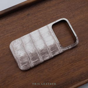 May include: A rectangular phone case featuring a light grey and white textured surface with a repeating crocodile skin pattern. The case has a large, open cutout at the top for a camera module. The text TRIS LEATHER is printed at the bottom.
