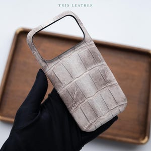 Op de afbeelding: Een lichtgrijze telefoonhoes met een gestructureerd krokodillenleerpatroon en een grote rechthoekige uitsparing voor de camera. Bovenaan de hoes staat de tekst TRIS LEATHER in kleine zwarte letters. De hoes heeft een rechthoekige vorm.