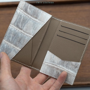 May include: A bi-fold wallet featuring a taupe textured interior with three horizontal card slots and two diagonal pockets accented with a light grey and white crocodile-embossed pattern. The text TRIS LEATHER is printed at the top centre.