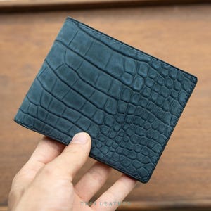 Könnte beinhalten: Ein dunkelblaues Bifold-Portemonnaie mit einer strukturierten Krokodilleder-Optik auf der Außenfläche. Das Portemonnaie hat eine matte Oberfläche und sichtbare Nähte an den Kanten. Der Text TRIS LEATHER ist unten mittig aufgedruckt.