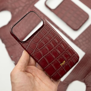 Op de afbeelding: Een diepbordeauxrode smartphonehoes met een gestructureerd krokodillenleerpatroon en de goudkleurige initialen YMP onderaan. De hoes heeft een grote rechthoekige uitsparing voor de cameramodule en is voorzien van de tekst SU LEATHER.