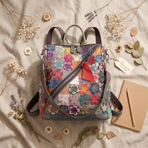 Könnte beinhalten: Ein mehrfarbiger Lederrucksack mit einem Patchwork-Gitterdesign, das mit zahlreichen kleinen, erhabenen Blumenapplikationen in verschiedenen Farben verziert ist. Die Tasche verfügt über einen diagonalen Reißverschluss, einen Tragegriff, verstellbare Schultergurte und einen roten Lederanhänger.