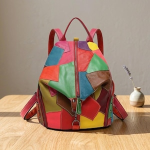 Könnte beinhalten: Ein mehrfarbiger Lederrucksack mit einem Patchwork-Design aus roten, grünen, blauen, gelben, violetten und braunen Feldern. Der Rucksack verfügt über einen zentralen vertikalen Reißverschluss, einen Tragegriff, zwei verstellbare Schultergurte und einen kleinen Metallverschluss.