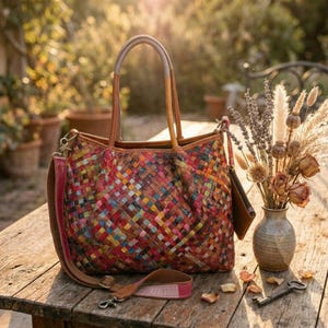 Puede incluir: Un bolso tote de cuero trenzado multicolor con un vibrante patrón de cuadros en tonos rojos, azules, amarillos y verdes. El bolso incluye dos asas altas de cuero, una correa de hombro desmontable y una pequeña bolsa de cuero a juego.