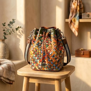 May include: A bucket bag featuring a multi-coloured woven leather pattern in shades of orange, blue, green, and yellow. The bag has a solid dark green leather top trim, a drawstring closure with two brown leather cords, and a detachable shoulder strap.