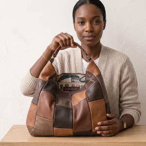 Puede incluir: Un bolso de estilo hobo confeccionado con un mosaico de paneles de cuero en tonos marrón, beige y chocolate oscuro. El bolso cuenta con un asa de cuero trenzado y un forro interior estampado con una escena de café y el texto LE CAFE DES ARTS.