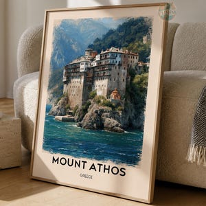 May include: A framed wall art print featuring a painting of a monastery perched on a rocky cliff overlooking blue sea water. The text MOUNT ATHOS GREECE is printed in black at the bottom of the off-white border surrounding the central landscape image.
