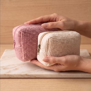 May include: Two rectangular zippered pouches with a textured, grid-like crochet pattern. One pouch is a dusty rose pink with a matching pink zipper, and the other is a cream off-white color featuring a metallic zipper with a brass-toned pull.