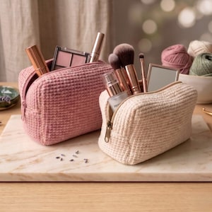 May include: Two rectangular zippered pouches with a textured waffle-knit exterior, one in dusty rose and one in cream. The pouches are filled with various cosmetic items, including makeup brushes, a small mirror, and tubes of beauty products.