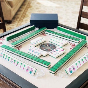 May include: A Mahjong set featuring white tiles with green backs and colorful floral illustrations arranged on a square table. The set includes four clear plastic tile racks, a central circular game component, and a dark blue storage box.