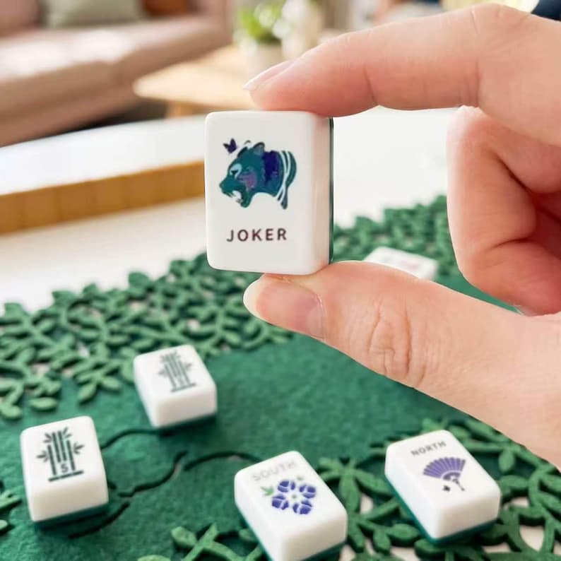May include: A white rectangular mahjong tile held between fingers, featuring a blue and green graphic of a creature and the text JOKER. Other tiles with bamboo, flower, and directional symbols are scattered on a green patterned surface.