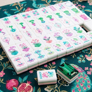 May include: A set of white mahjong tiles with green and pink floral, lantern, and character designs. The tiles feature letters N, E, W, S, and the word JOKER. One tile is separate, and a translucent green rectangular block is included.