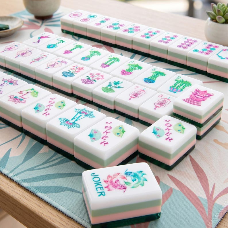 May include: A set of rectangular mahjong tiles featuring white tops with colorful floral, bamboo, lantern, and fish illustrations, along with the text Joker and W. Each tile has a layered design with a white top, a light green middle, and a dark green base.