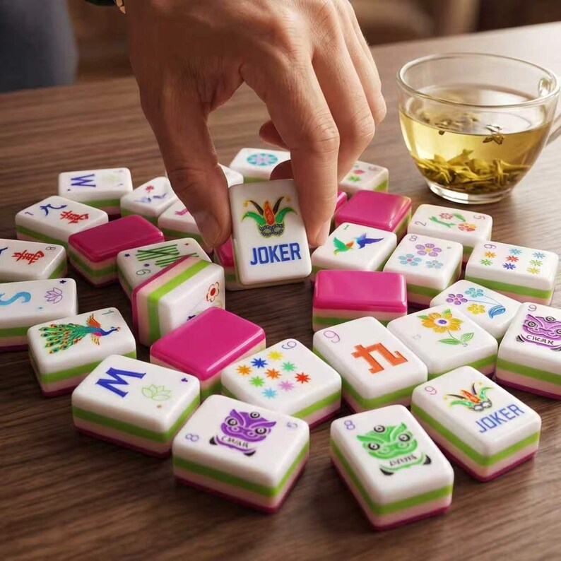 May include: A collection of rectangular mahjong tiles with white faces featuring colorful illustrations of flowers, birds, dragons, and the word JOKER. The tiles have pink and green layered bases and are scattered across a wooden surface.