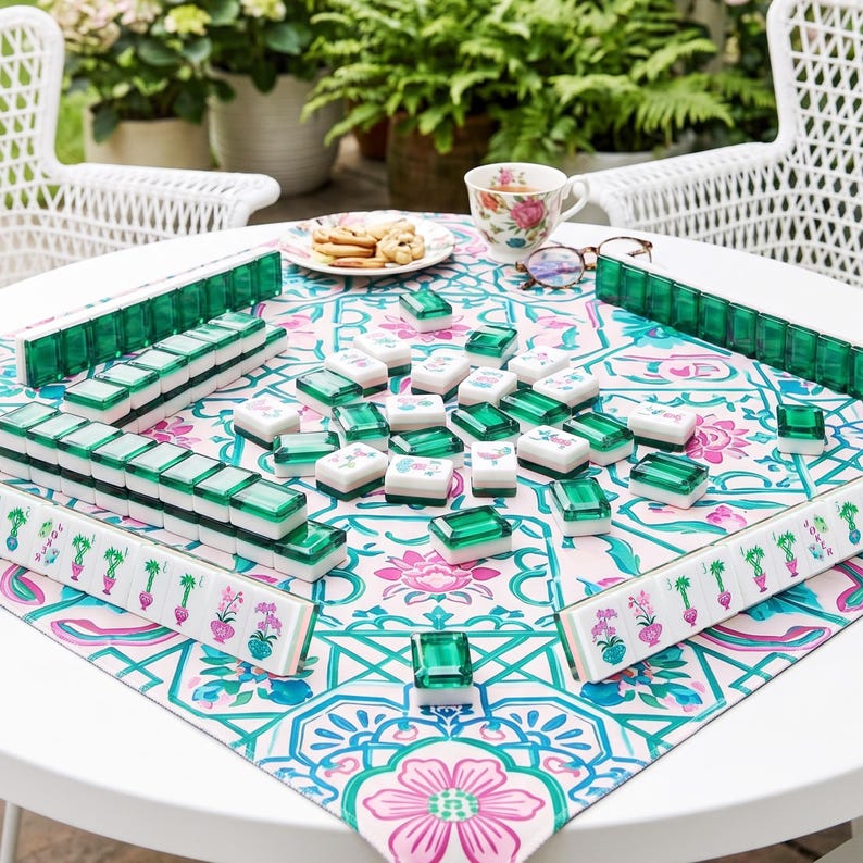 May include: A Mahjong set featuring white tiles with green translucent backs and colorful floral or botanical illustrations on the faces. The tiles are arranged on a square tablecloth with a pink, green, and white floral pattern on a white table.