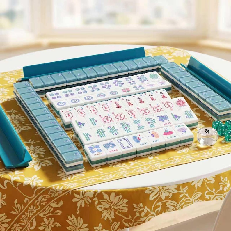 May include: A Mahjong set featuring white tiles with colorful illustrations of flowers, fans, and Chinese characters, arranged in rows. The set includes teal tile racks and a clear die. The tiles display various symbols, numbers, and the word JOKER.
