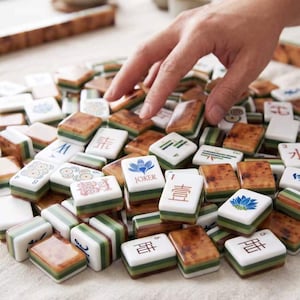 May include: A collection of rectangular Mahjong tiles featuring white faces with various colorful symbols, Chinese characters, and a blue lotus flower labeled JOKER. The tiles have layered green and brown sides with a polished, smooth finish.