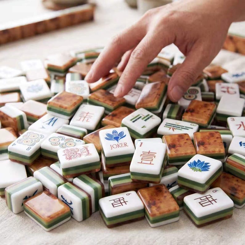 May include: A collection of rectangular Mahjong tiles featuring white faces with various colorful symbols, Chinese characters, and a blue lotus flower labeled JOKER. The tiles have layered green and brown sides with a polished, smooth finish.