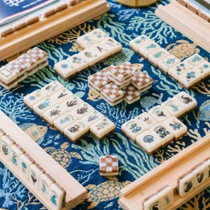 May include: A mahjong set featuring off-white tiles with various botanical and marine illustrations, alongside brown and white checkered tiles. The tiles are arranged on a blue fabric mat with a gold-colored sea turtle and coral pattern.