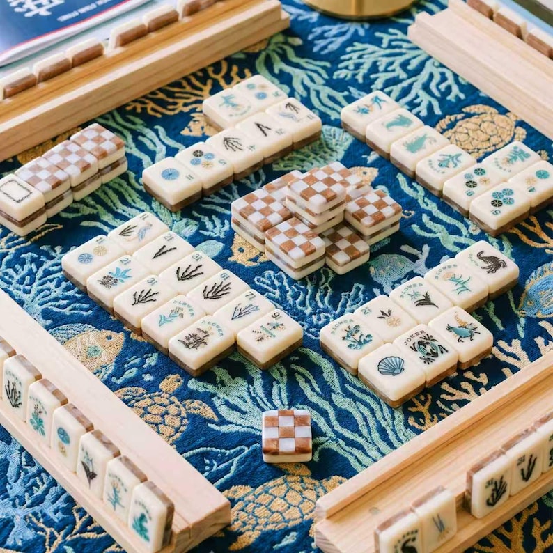 May include: A mahjong set featuring off-white tiles with various botanical and marine illustrations, alongside brown and white checkered tiles. The tiles are arranged on a blue fabric mat with a gold-colored sea turtle and coral pattern.
