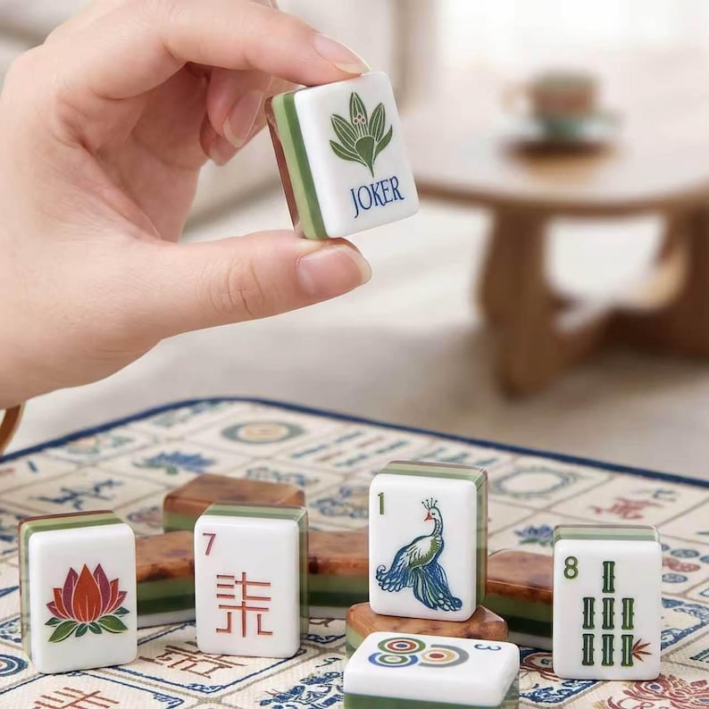May include: A white and green Mahjong tile featuring a green lotus-like graphic and the word JOKER in blue text. The tile has a layered construction with a white top surface and a translucent green base, displaying intricate game symbols.