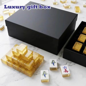 May include: A large black rectangular box labeled Luxury gift box, accompanied by a collection of translucent gold glitter mahjong tiles. Some tiles are stacked in a pyramid, while others are placed individually, featuring blue and red ink designs.