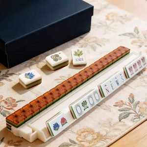 May include: A Mahjong set featuring white tiles with green and brown accents, including tiles with floral designs, the word NEWS, and a JOKER tile. The set includes a white tile rack and a black storage box, displayed on a floral patterned surface.