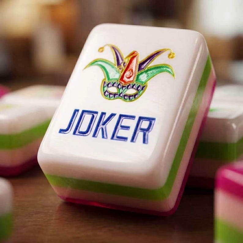 May include: A white rectangular mahjong tile featuring a colorful jester mask graphic in purple, green, and red, with the word JOKER embossed in blue capital letters. The tile has layered side panels in green and pink, with a smooth, glossy finish.