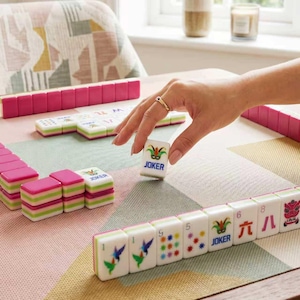 May include: A set of Mahjong tiles featuring pink, green, and white color schemes with various illustrations including hummingbirds, floral patterns, and Joker cards. The tiles are arranged in rows and stacks on a patterned mat with one tile held.