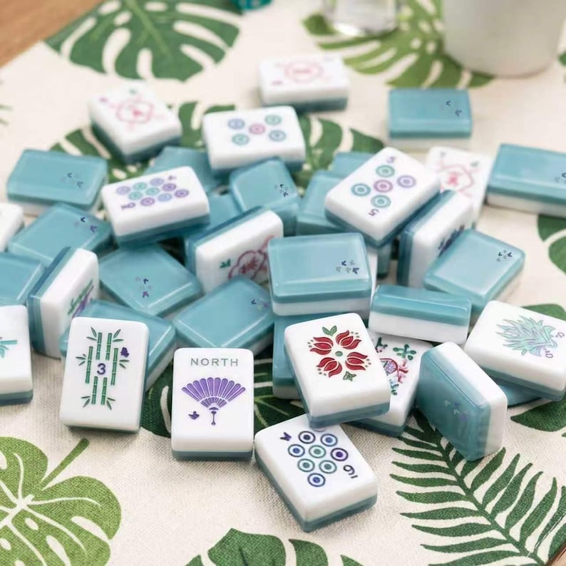 May include: A collection of rectangular mahjong tiles with white faces and teal backs. The tiles feature various colorful engraved designs including a purple fan labeled NORTH, a green bamboo stalk, a red flower, and circular patterns in teal and purple.