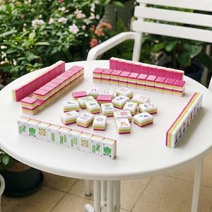 May include: A Mahjong set featuring white tiles with colorful graphic illustrations and pink backs. The set includes several rows of stacked tiles and individual tiles arranged on a white table, displaying various symbols, flowers, and a Joker tile.